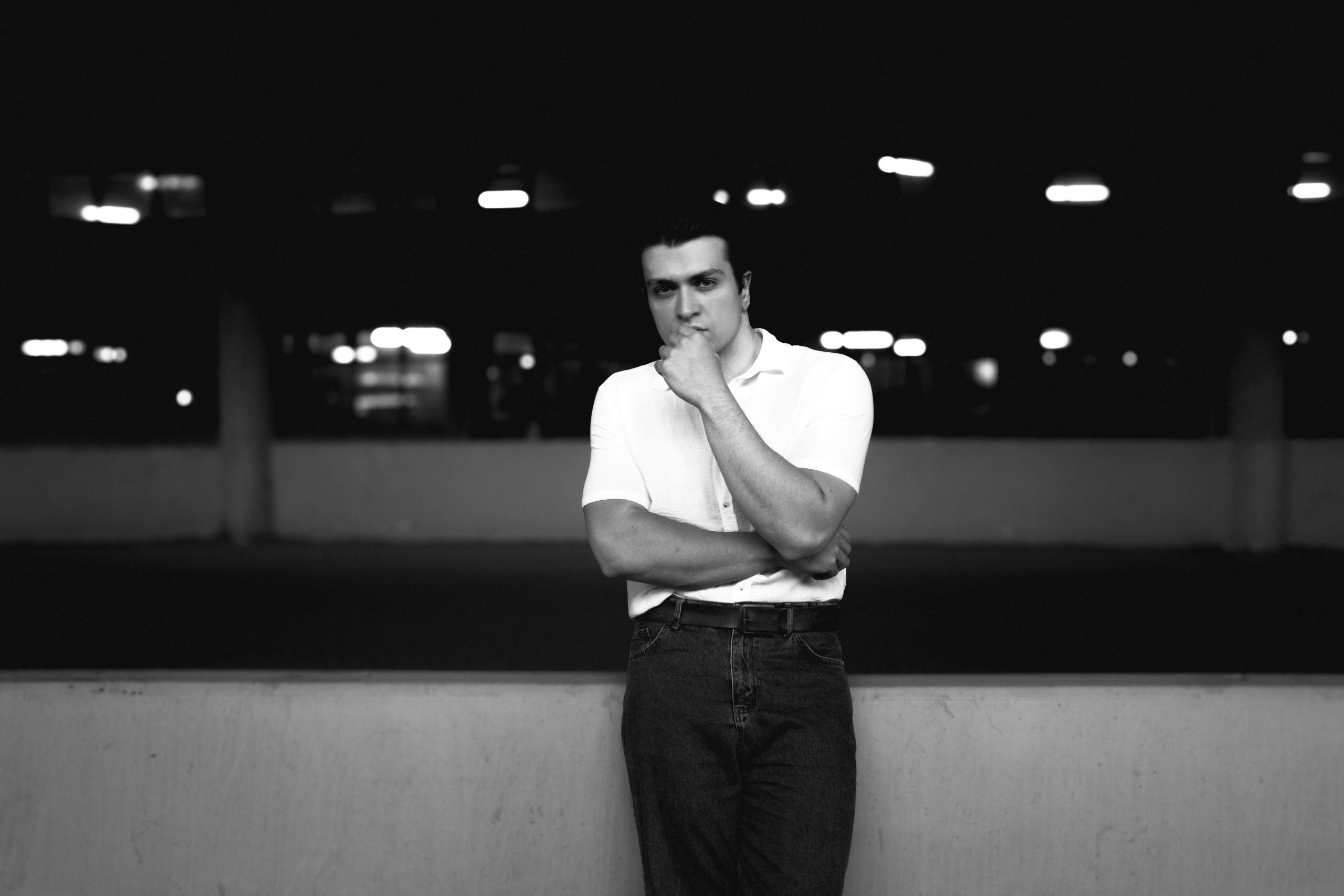 A black-and-white photo of a man in a thoughtful pose, leaning against a low wall in an urban setting at night. He is wearing a white shirt and jeans, with blurred lights in the background.